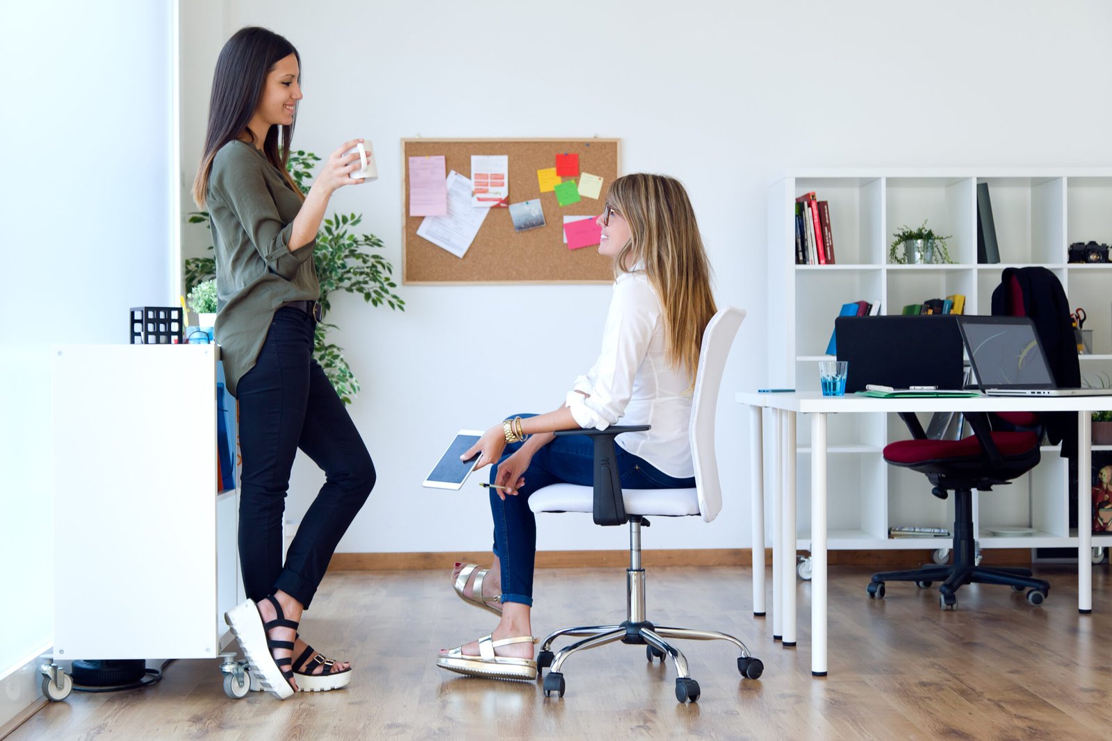 Two businesswomen discussing work in a modern office, one standing with a coffee mug and the other sitting on a white office chair holding a tablet.